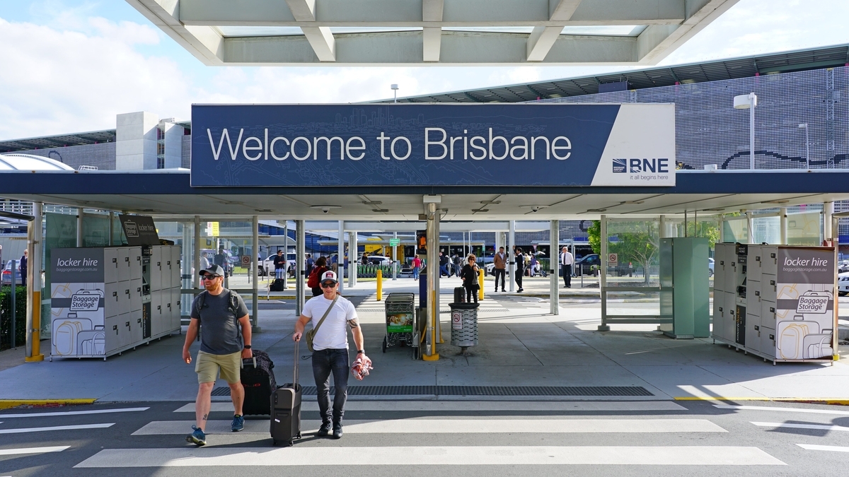 「Welcome to Brisbane」と書かれた大きな看板の下の横断歩道を、スーツケースを持った二人の男性が歩いている様子