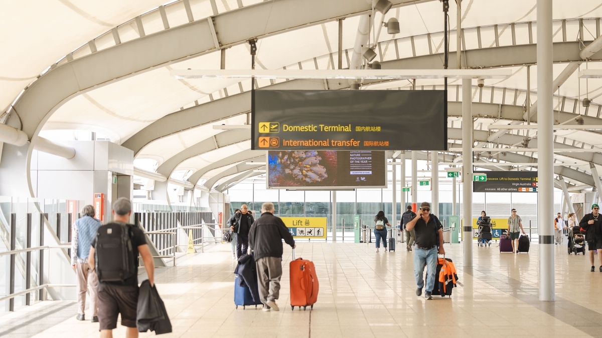 「Domestic Terminal」等の案内板が吊り下げられた空港内で、キャリーケースを持った多くの人々が行き交う通路の風景