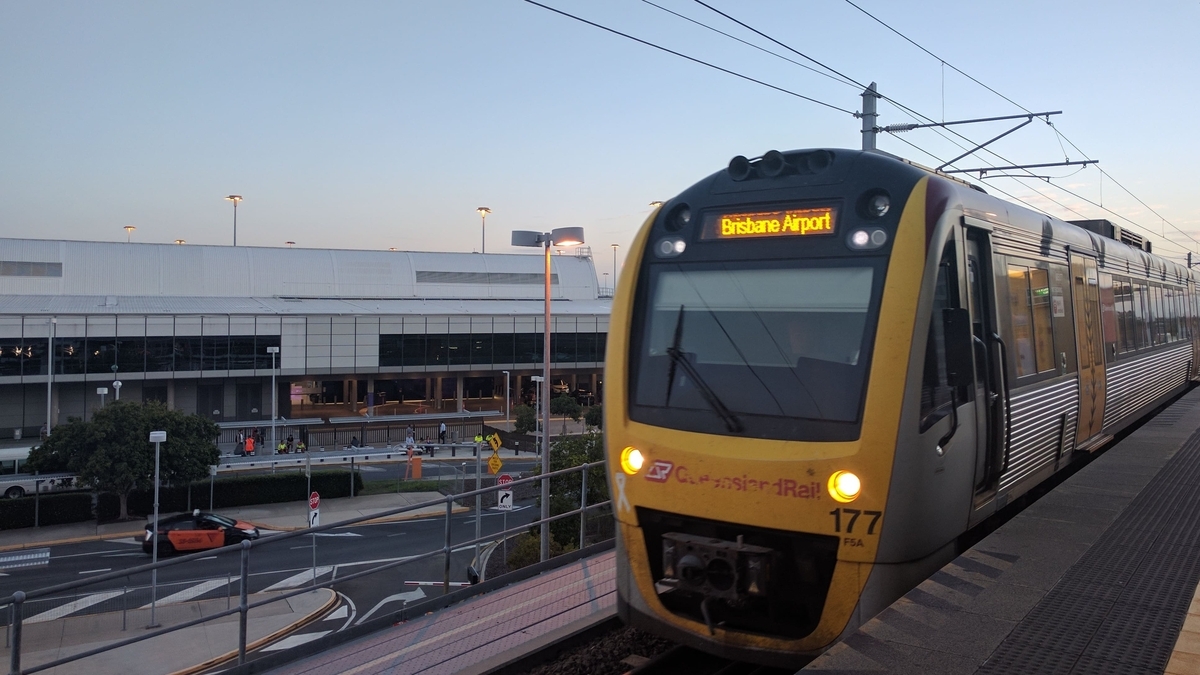 電光掲示板に「Brisbane Airport」と表示された黄色と銀色の電車が、プラットホームに入線している様子