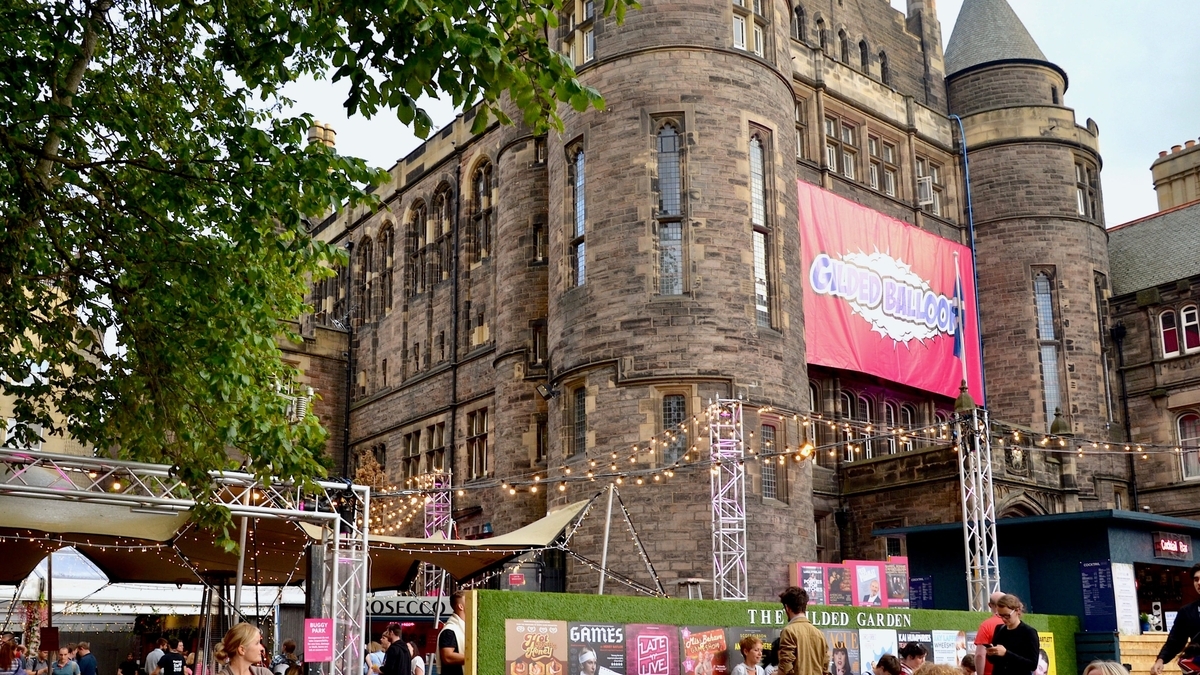 石造りの歴史的な建物の壁面に「GILDED BALLOON」というピンク色の大きな看板が掲げられている様子。手前の広場にはテントやストリングライトが設置され、多くの人々が集まっている