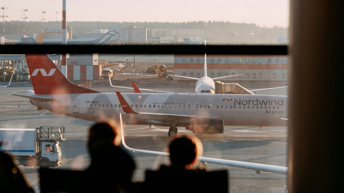 空港の窓ガラス越しに、白地に赤い尾翼の「Nordwind Airlines」の機体が駐機場に停まっている様子。手前には外を眺める人々の頭から肩にかけてのシルエットが黒く写り込んでいる