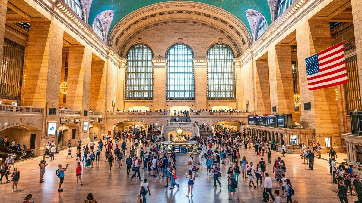 高いアーチ型の天井と大きな窓がある広大な駅のコンコースの風景です。中央にある時計台の周りを大勢の人が行き交い、右側の天井からは大きなアメリカ国旗が吊るされています。