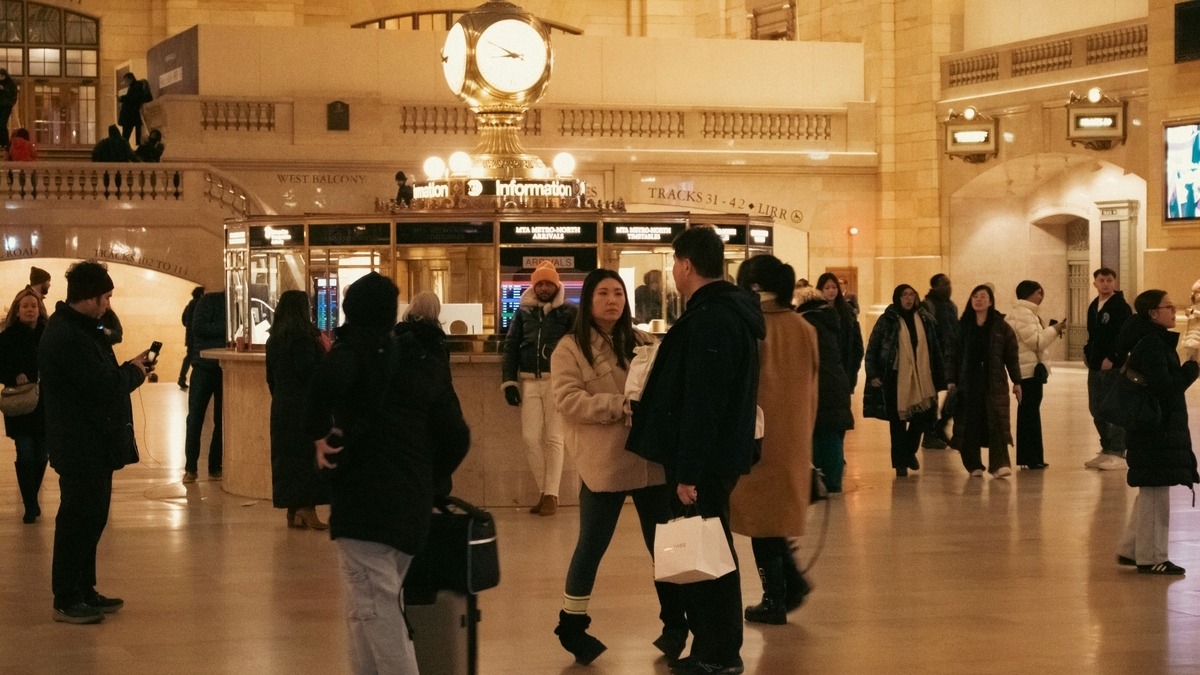 駅のコンコースの中央にある、四面時計が乗った丸いインフォメーションブースの様子です。コートなどの冬服を着た多くの人々が周囲を行き交う中、手前で数人が立ち止まっています