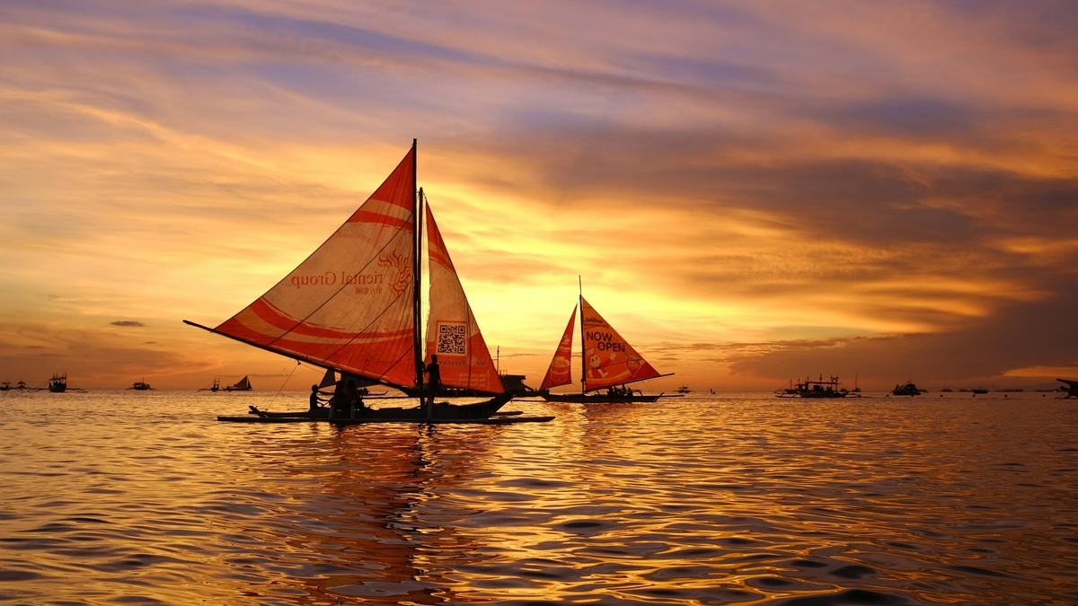オレンジ色に染まる美しい夕焼け空と、その光を反射する海面の風景です。水面には特徴的な帆を持つ複数のボートが浮かび、船上には人々のシルエットが見えます