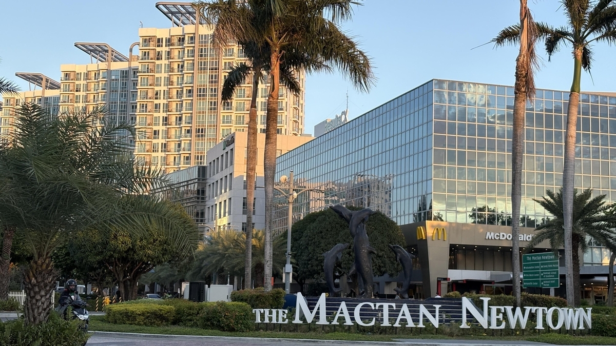 「THE MACTAN NEWTOWN」という白い立体文字の看板とイルカのオブジェ。背景にはヤシの木が立ち並び、マクドナルドの店舗が入るガラス張りのビルや近代的な高層マンションが建っています