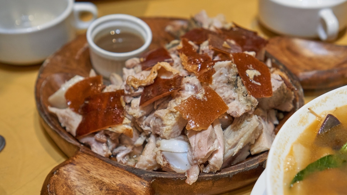 木の皿に山盛りになった、皮がパリッと焼かれた豚肉のロースト。傍らには茶色いソースが入った小さな器が置かれ、右端には野菜が入ったスープの一部が写っています