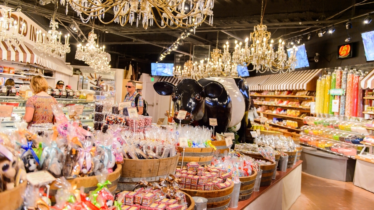 多数のシャンデリアが吊るされた菓子店内の様子。中央には大きな白黒の牛のオブジェが置かれ、木樽や棚に色鮮やかなキャンディなどのスイーツが大量に陳列される中、客が買い物をしています。