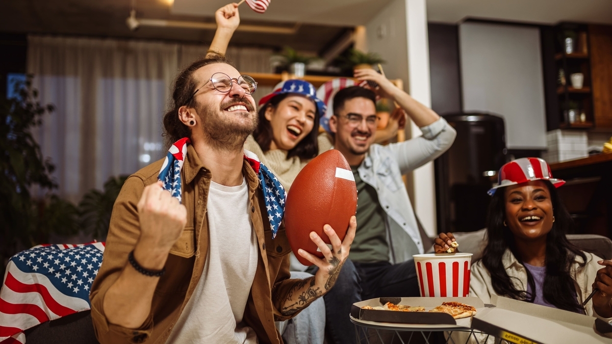室内でアメリカ国旗柄の帽子やバンダナを身につけた4人の男女が、アメリカンフットボールのボールを手に歓声を上げています。手前のテーブルにはピザやポップコーンが置かれています。