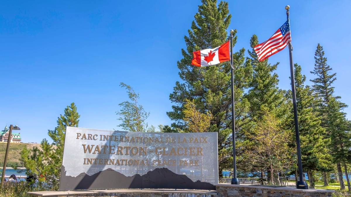 「WATERTON - GLACIER INTERNATIONAL PEACE PARK」の文字と山のシルエットが切り抜かれた金属製の看板。背後には針葉樹が茂り、カナダとアメリカの国旗が並んで風にたなびいています。