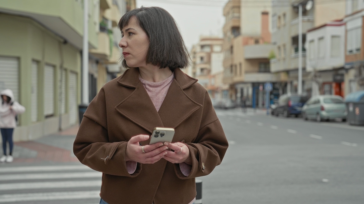 茶色いショートコートを着た黒髪の女性が、両手でスマートフォンを持ちながら横断歩道付近の路上に立っています。女性は顔を右に向けており、背景にはぼやけた街並みと駐車された車が写っています。