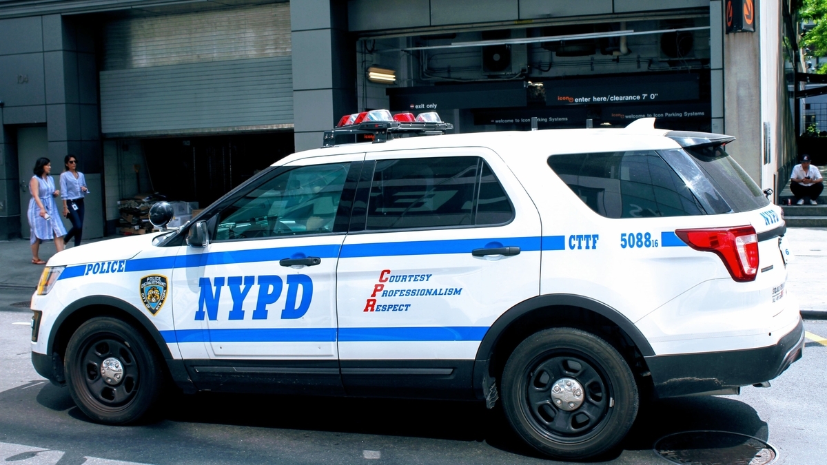 街路に停車している、白地に青いラインと「NYPD」の青い文字が入った警察のSUV車両です。車両の屋根には赤のパトランプがあり、背景にはビルの入り口や数人の歩行者が写っています。