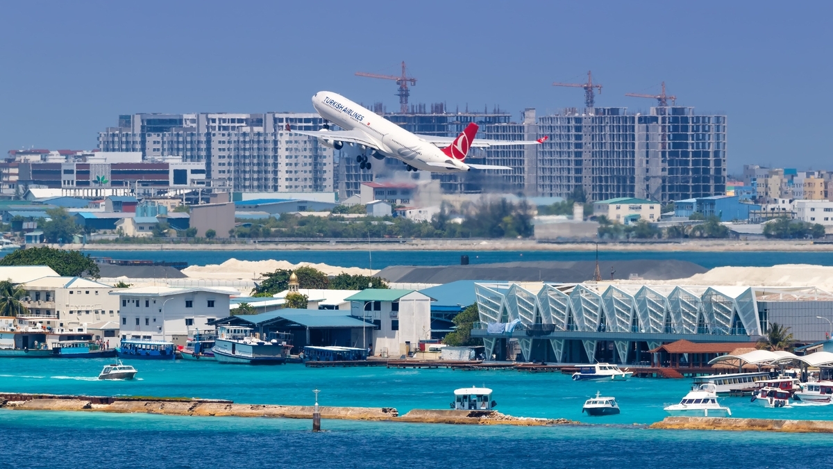 青い海と建造物が広がる沿岸で、赤い尾翼の大型旅客機がクレーンの立ち並ぶ市街地を背景に離陸している光景。