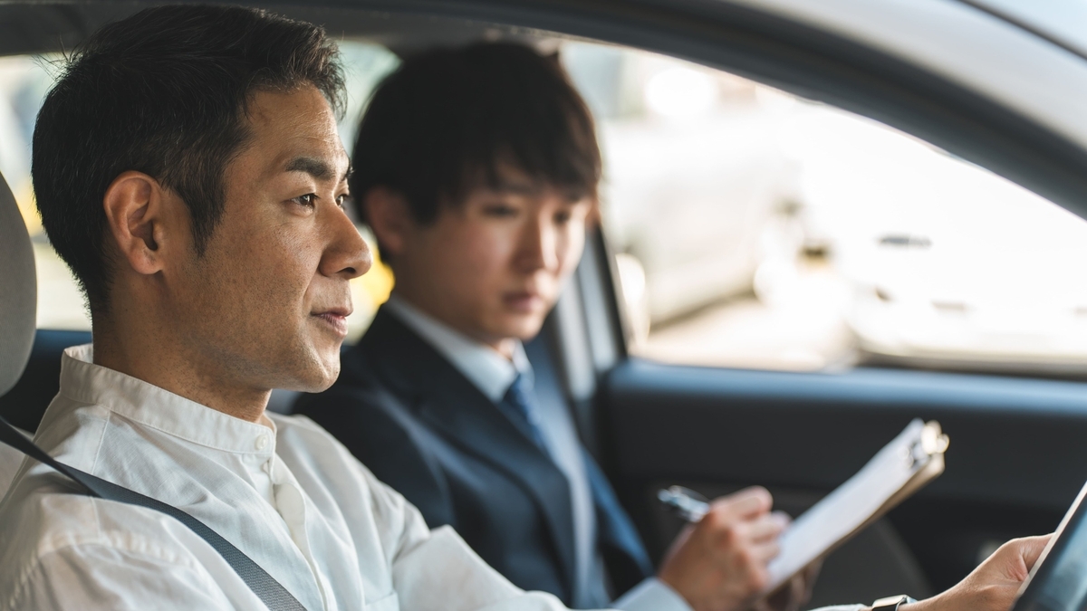 車の運転席で前方を見つめる白いシャツの男性と、助手席でバインダーにメモを取るスーツ姿の男性