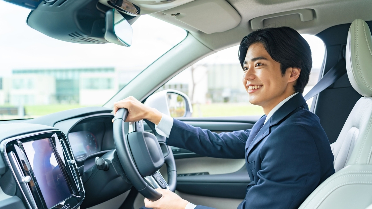 スーツを着て車の運転席に座り、ハンドルを握りながら笑顔で進行方向を見つめる男性。