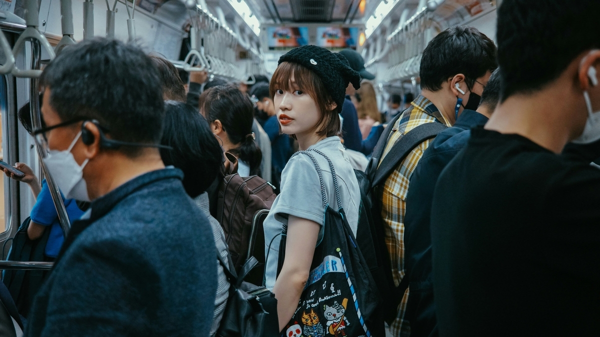 つり革が並ぶ混雑した電車内で、黒いニット帽をかぶりバッグを肩に掛けた女性が後方を振り返っている。