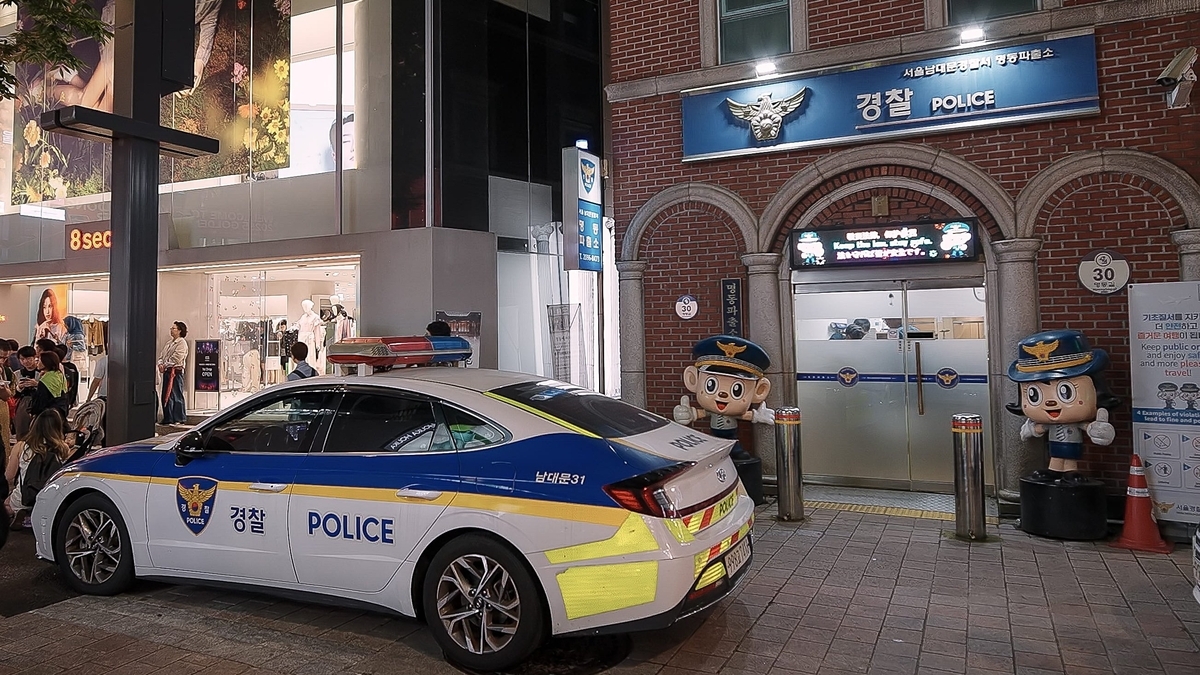 「POLICE」と書かれた車両が、赤レンガ造りの警察施設と制服姿のキャラクター像の前に止まっている。