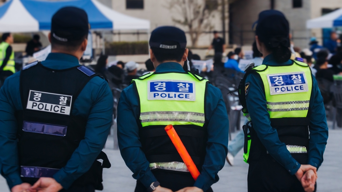 「POLICE」と表示された反射ベストと濃紺の制服を着た3人の警察官が、人々の前に背を向けて立っている。