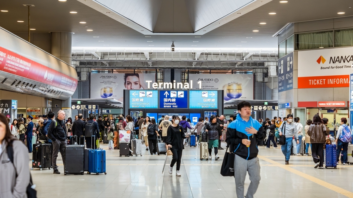 スーツケースを持った多くの旅行者が行き交う空港ターミナル内の広いコンコースで、中央には「Terminal 1」と書かれた国際線や国内線の青い案内表示板が設置されています。   