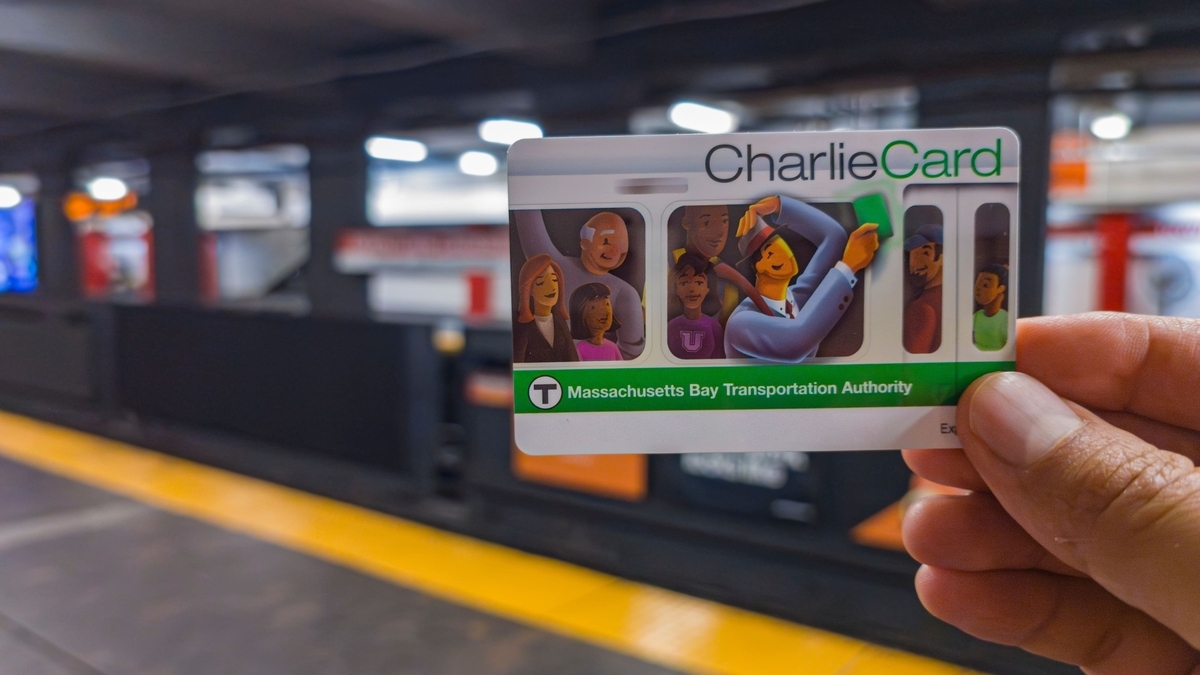 ぼやけた地下鉄のホームを背景に、人物のイラストが描かれた「CharlieCard」という緑と白の乗車カードを手で持っている様子が写されています。
