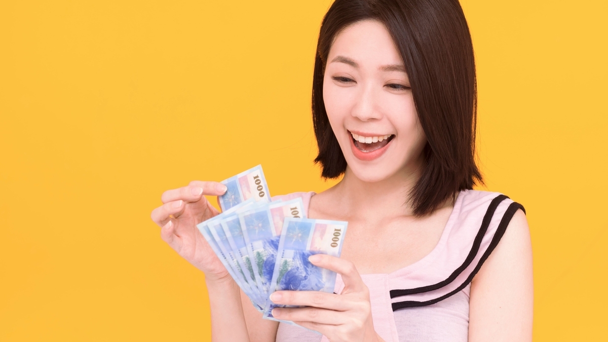 黄色い背景の前で、複数枚の青い紙幣を両手に持ち、口を開けてうれしそうな笑顔を見せている女性の様子です。手にしている紙幣には「1000」などの数字が印刷されています。