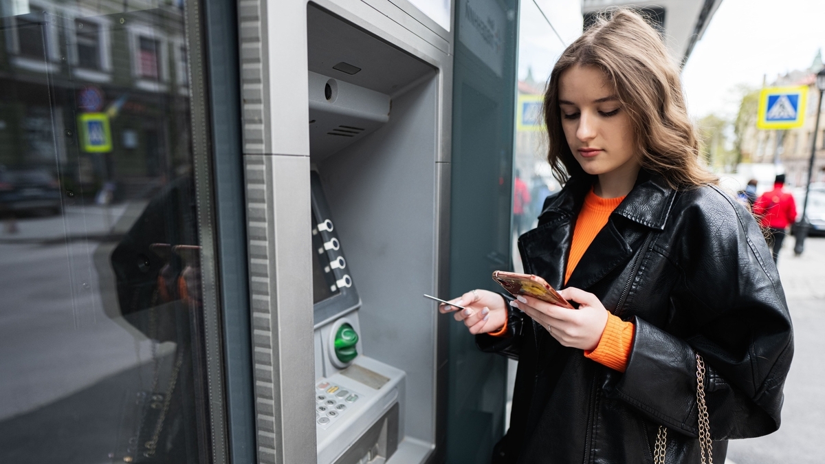 屋外に設置されたATMの前に立つ、黒いレザージャケットを着た女性の様子です。右手でスマートフォンの画面を見ながら操作し、左手にはカードを持っています。