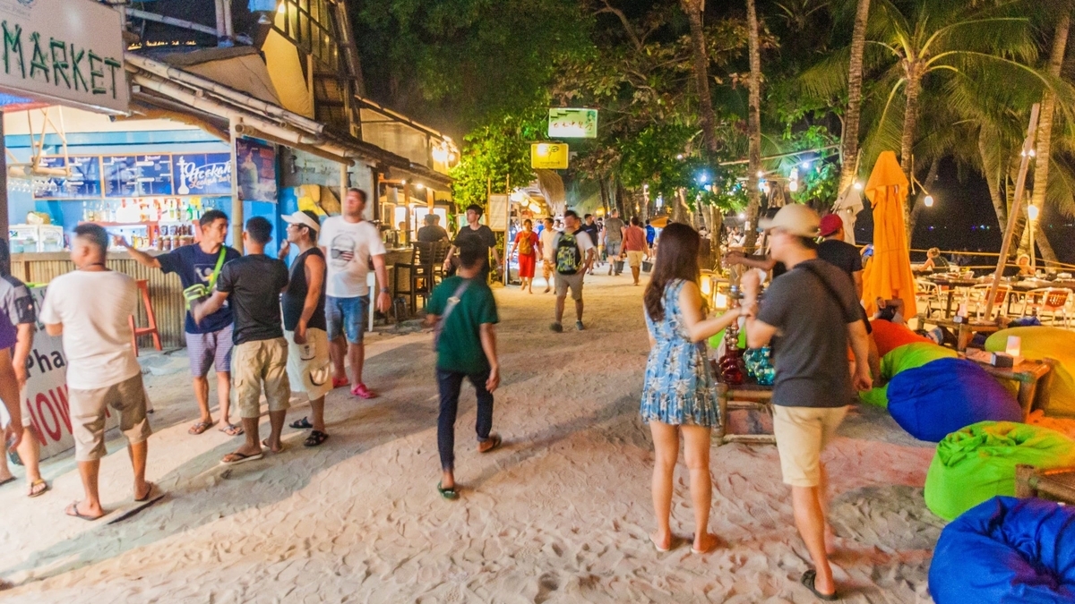 夜間の砂地の通路に多くの人々が行き交う様子です。左側には「MARKET」と書かれた看板を掲げる青い外観の店舗が並び、右側には緑や青のクッションが置かれた席が設けられています。   