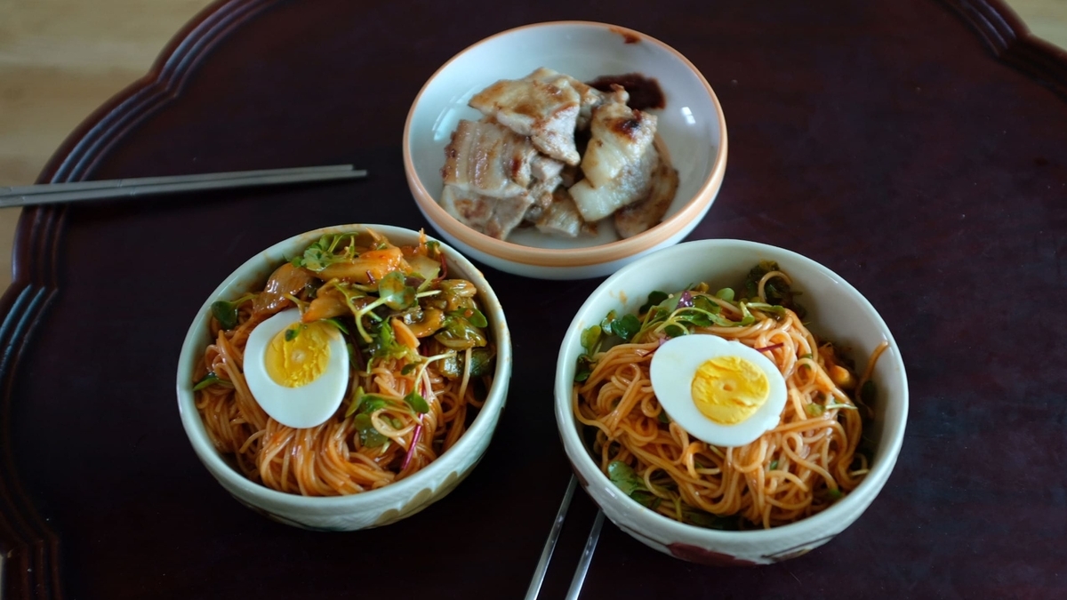 茶色い円形のテーブルに、ゆで卵や野菜が乗った赤い麺料理の丼が2つ並んでいます。奥には、タレがかかった厚切りの豚肉を焼いた料理が白い小皿に盛られ、左手前には銀色の箸が置かれています。   