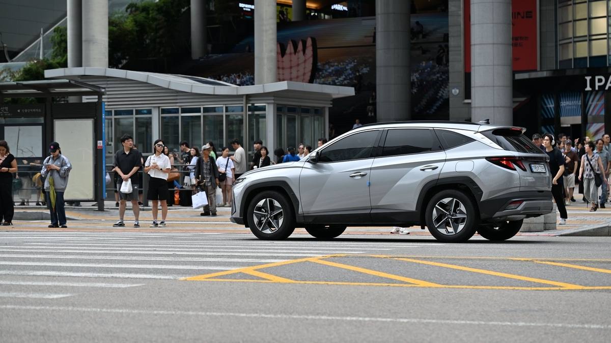 近代的なビルが建つ市街地の交差点で、シルバーのSUV車が車道を走行しています。奥の横断歩道やガラス張りのバス停付近には、信号待ちをしている複数の歩行者の姿が見えます。   