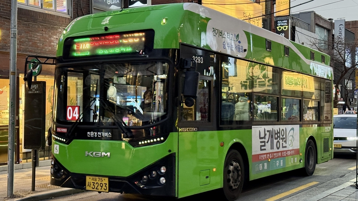 夕暮れ時の市街地を走行する、鮮やかな緑色の路線バスです。車体の前面には「04」と書かれた赤い標識や電光掲示板があり、側面には病院の広告や韓国語の文字が描かれています。   