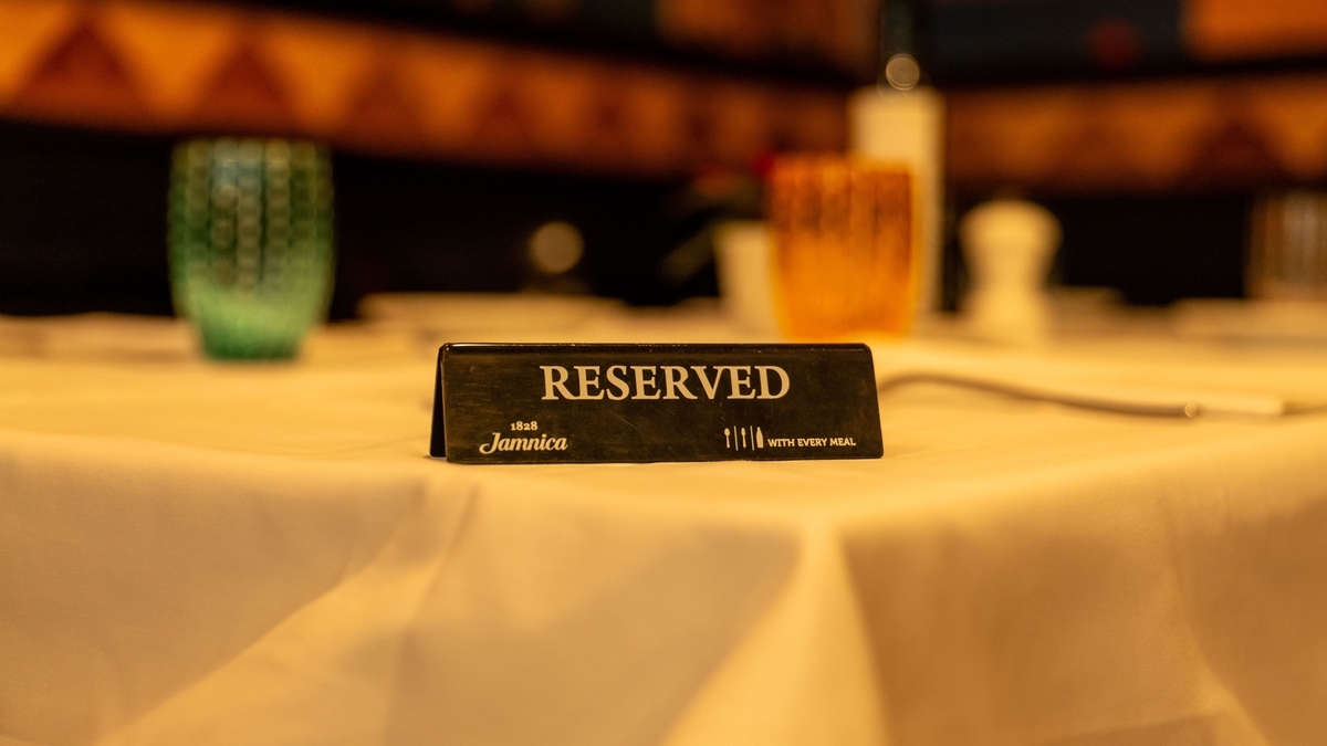 白いテーブルクロスが敷かれたテーブルの中央に、「RESERVED」と白い文字で印字された黒い卓上プレートが置かれています。奥にはピントがぼやけたグラスなどが配置されています。