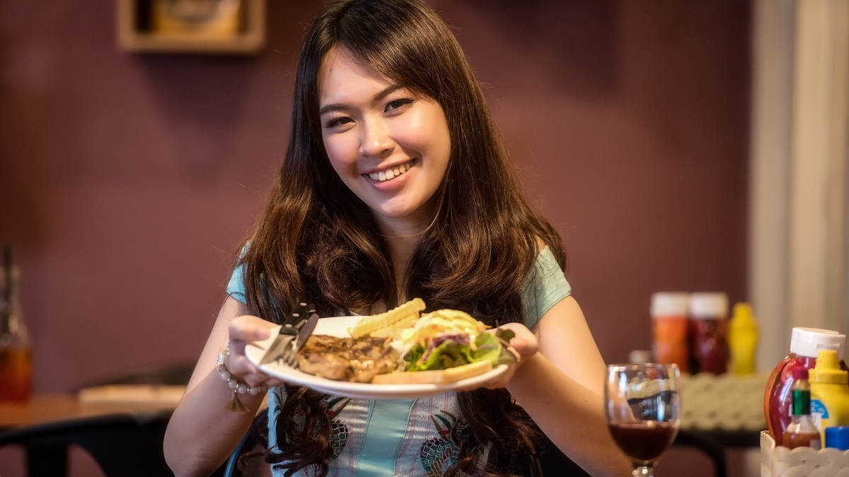長い髪の女性が笑顔で、焼いた肉、フライドポテト、サラダが盛り付けられた白い皿を両手で前に差し出しています。手前にはワイングラスやケチャップなどの調味料が置かれています。