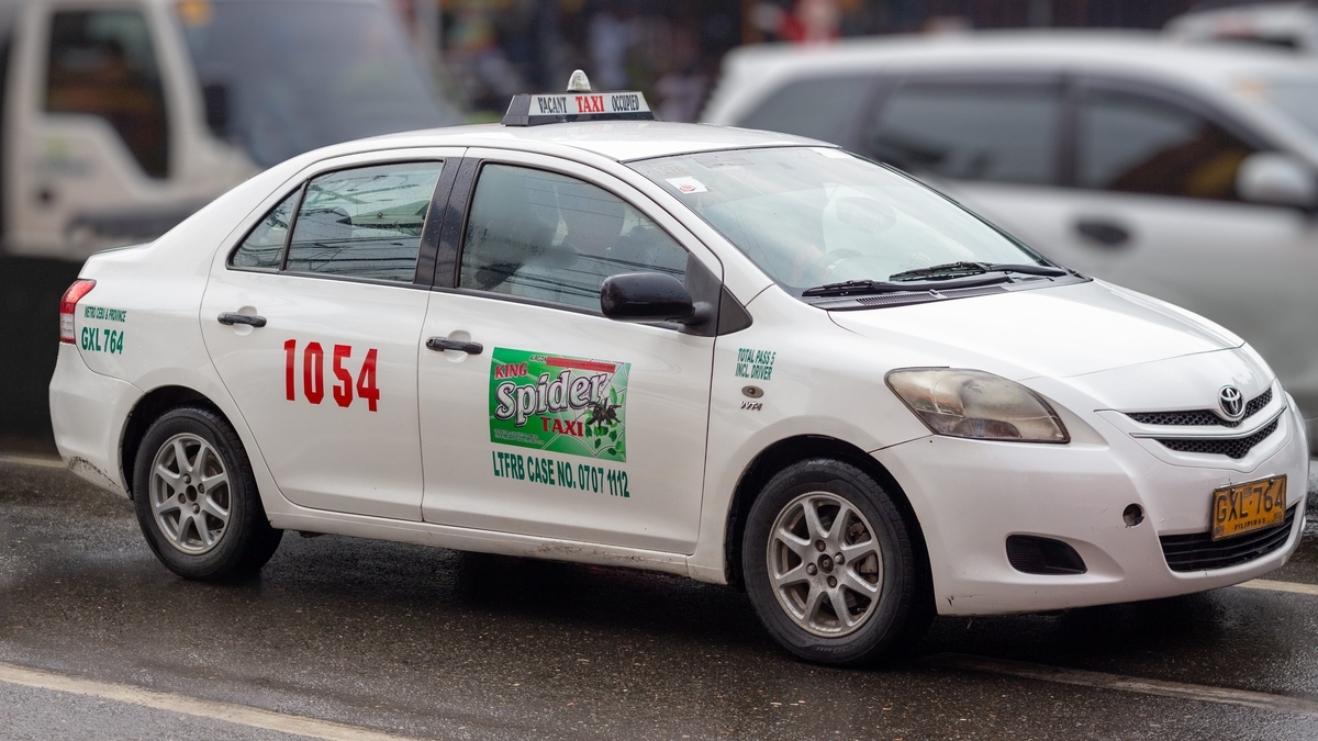 濡れた路面を走る白いタクシーの様子です。車体の側面には赤い数字の「1054」と緑の「Spider TAXI」のロゴが描かれ、屋根には行灯が設置されています。