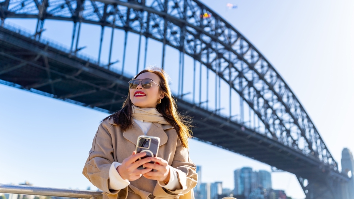 大きな金属製のアーチ橋を背景に、サングラスとコートを着用した女性が、スマートフォンを両手で持ちながら微笑んでいます。