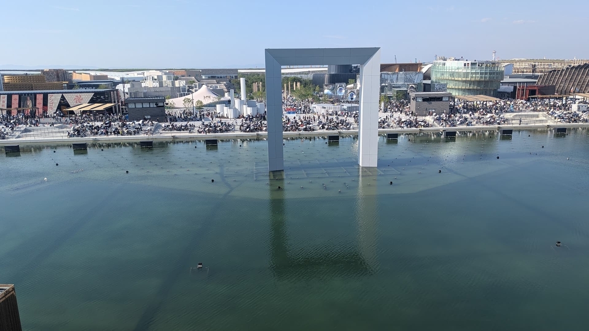 大きな水盤の中央に巨大な四角枠のモニュメントが立ち、対岸には多くの来場者と近未来的なパビリオン群が並ぶ様子を上から見下ろした光景