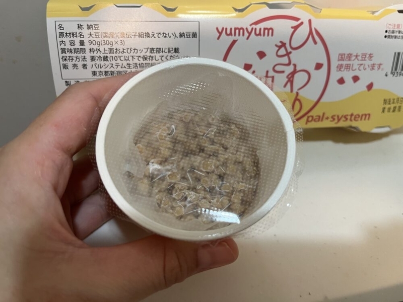片手で納豆のカップを持っている。納豆にはたれやからしがついておらず、フィルムがかかっている。