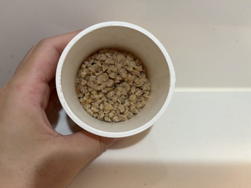 小さい納豆のカップを左手で持っている。中にはひきわり納豆が入っている。