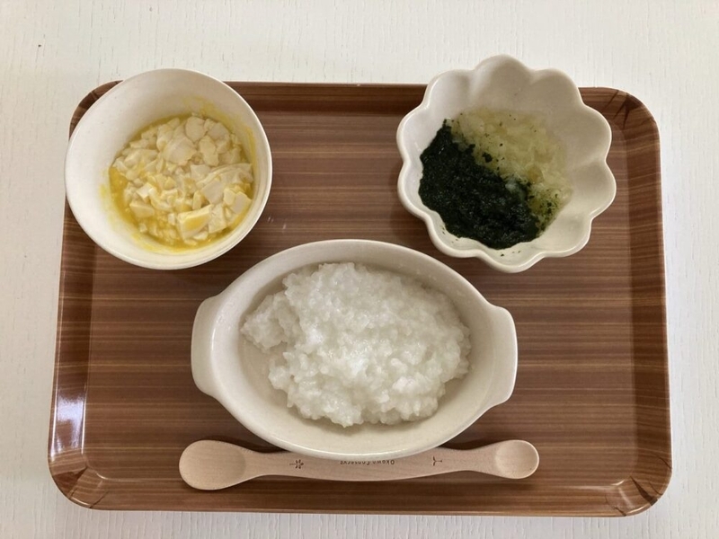 お盆の上に離乳食が置かれている。お粥、とうもろこしと豆腐の和物、ほうれん草のサラダ。手前にスプーンが置いてある。