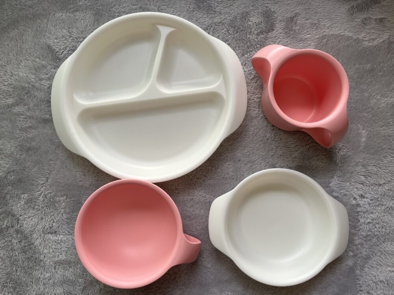 床に、プラスチックの大皿、コップ、お椀、小皿が並べられている。大皿と小皿は白、コップとお椀はピンク色である。