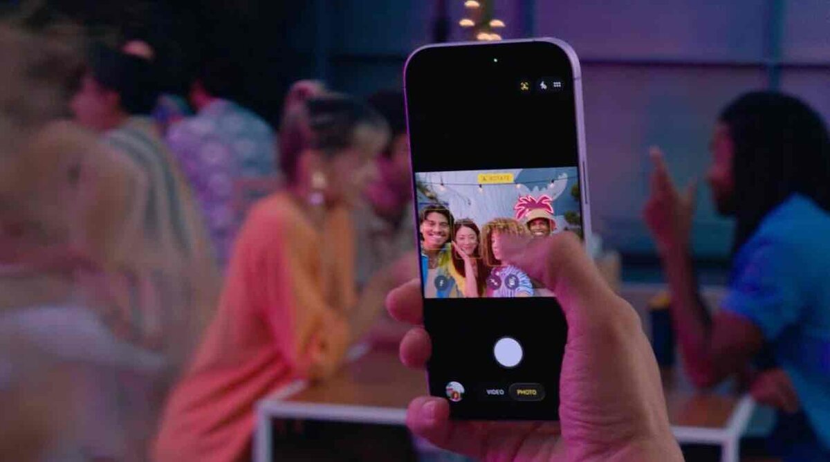 夜の屋外テラスでiPhoneを手に自撮りを撮影している様子。画面には4人の笑顔が映り、背景には暖色の照明と人々の会話がぼんやりと写っている。