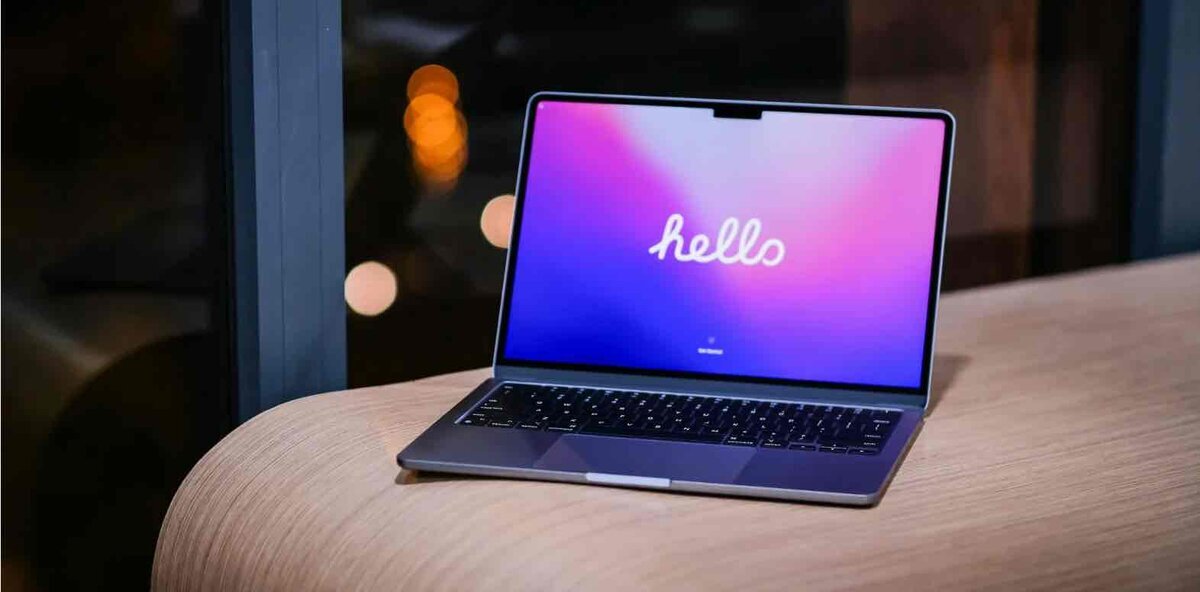 夜の室内で木のテーブルの上に置かれたMacBookが、紫からピンクのグラデーション背景に「hello」と表示した画面を開いている様子