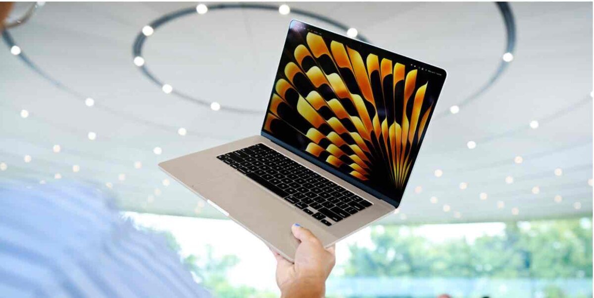 屋内の明るい空間で、人物が片手で持ち上げたMacBookが開かれており、画面には黄色と黒の抽象的なパターンが大きく表示されている様子