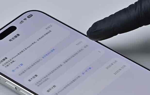 iPhoneの設定画面でバッテリー情報（最大容量や状態）を確認している様子。黒い手袋をした指が画面を指している