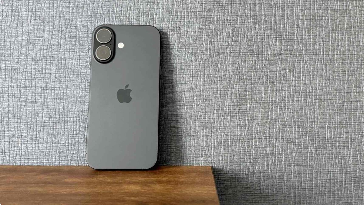 木製のデスクに立てかけられた、iPhone 16(ブラック)の背面。垂直に並んだ2つのカメラレンズとAppleロゴが特徴的な、落ち着いた質感のクローズアップ写真