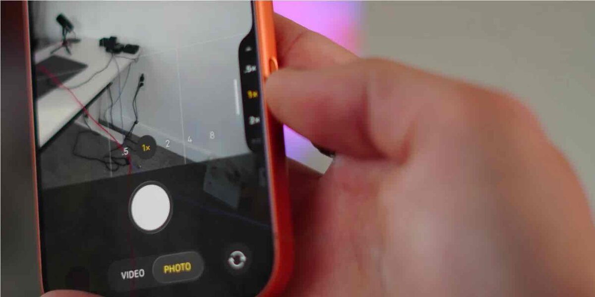 iPhoneのカメラアプリを起動した状態で、側面のカメラコントロール（ボタン）を指で操作している様子。画面には撮影倍率やシャッターボタンが表示され、実際の撮影操作を示すクローズアップ写真。