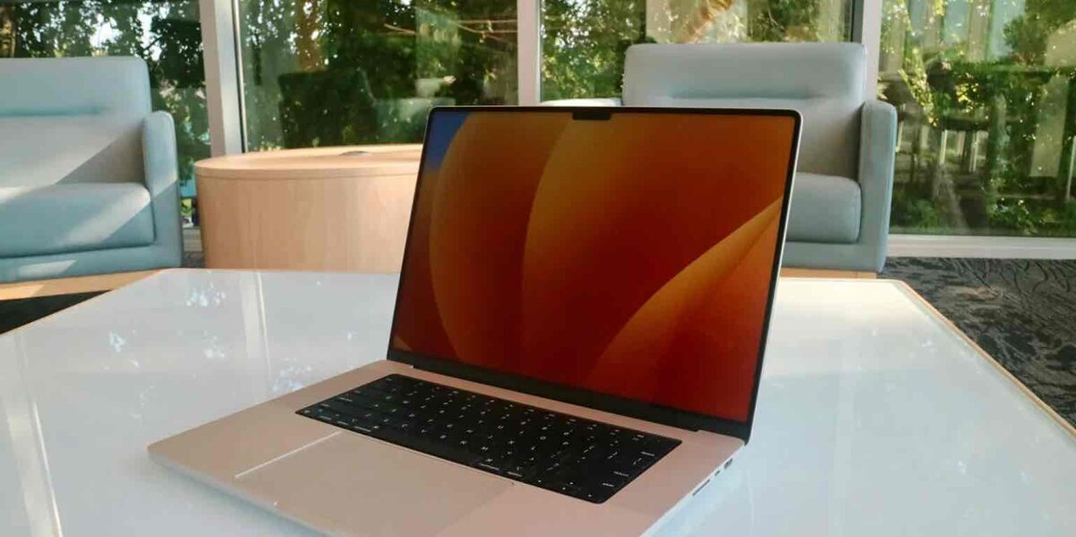 明るい窓際のラウンジにある白いテーブルの上に置かれた、シャンパンゴールド系のMacBookの斜め前からのショット。画面にはオレンジと黄色の抽象的なグラデーションの壁紙が表示されており、ベゼル上部にはノッチが見える。背景には水色のモダンな椅子と庭の緑がぼけて写っている