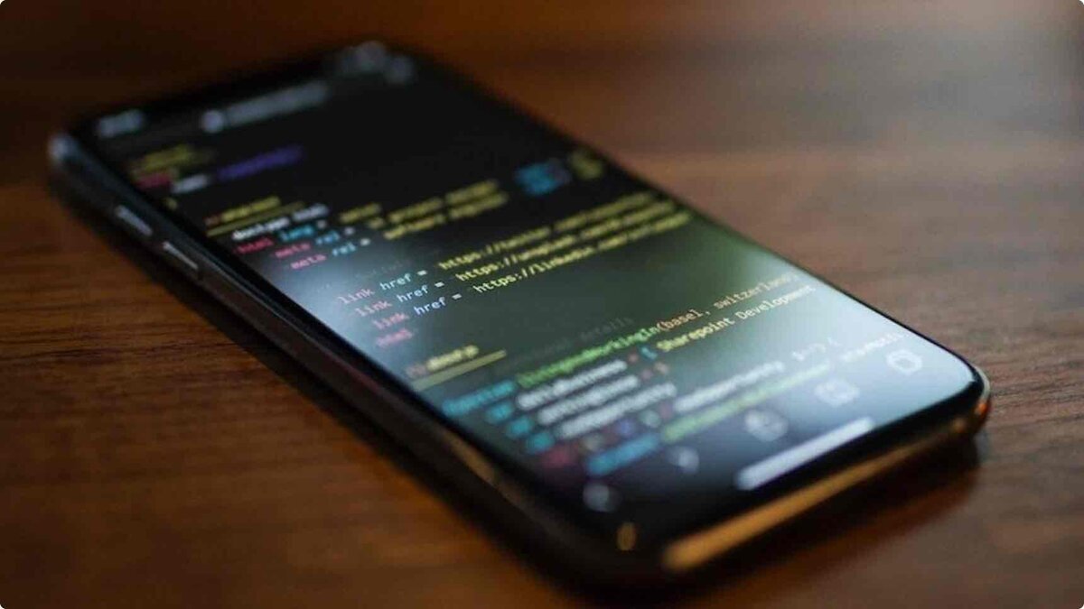 暗所において、iPhoneの画面上にHTMLコードのソースが表示されている様子。画面内にはソーシャルメディアのリンク(Twitter, LinkedIn等)を含む link href タグや、開発拠点の記述が並んでいる