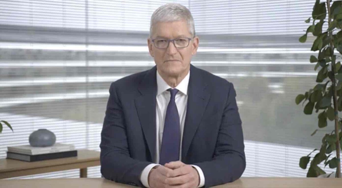 紺色のスーツとネクタイを着用し、眼鏡をかけたティム・クック(Apple CEO)が、デスクで両手を組み、正面を見据えて座っている様子。背景にはブラインド越しに光が差し込むオフィス