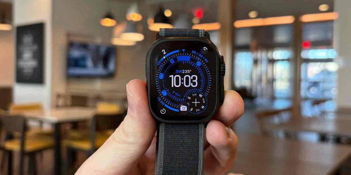 に持ったApple Watch Ultraのディスプレイにコンパス付きウォッチフェイスが表示され、時刻10:03と方位SW235°が確認できる様子。背景はカフェのような室内空間がぼかされている