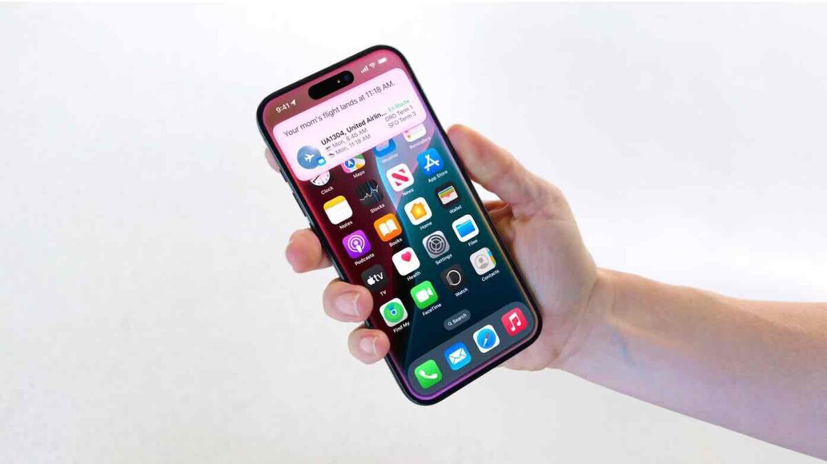 手に持ったiPhoneのホーム画面に、フライト情報のライブ通知が表示されている。白い背景の前で、Dynamic Island付近に航空便の到着案内が重なって見える