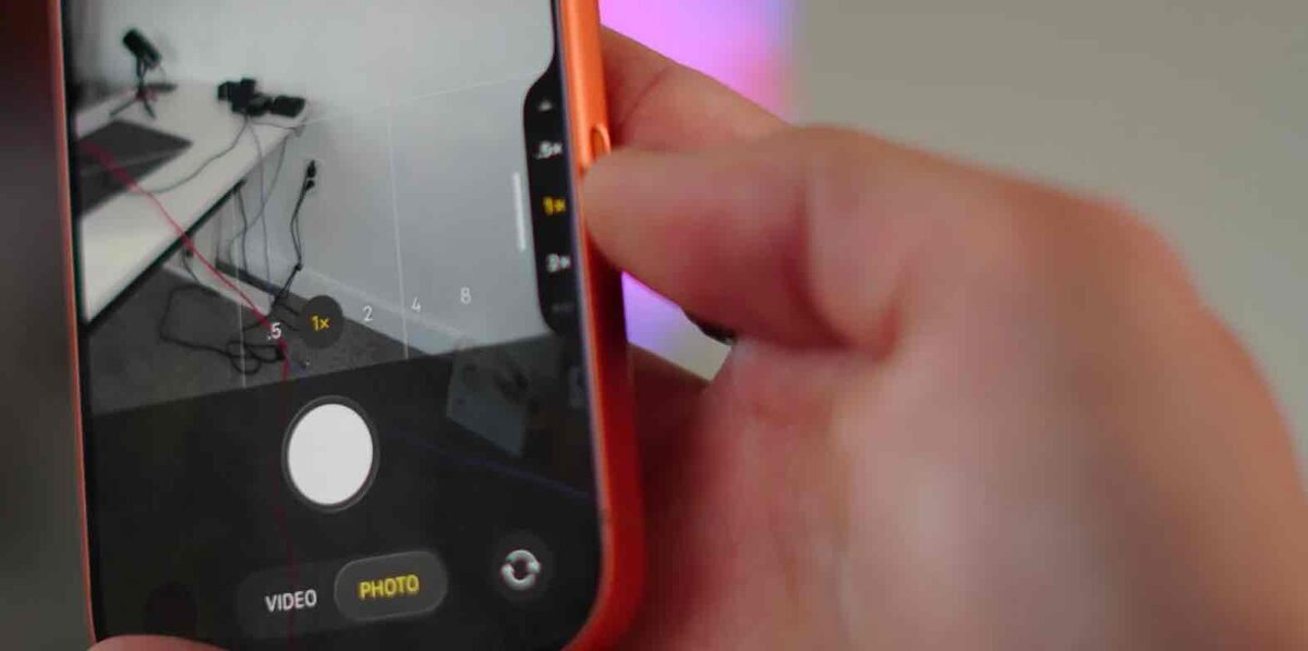iPhoneを手に持ち、カメラアプリのズーム操作画面を表示している様子。画面下にはシャッターボタンとPHOTOモードが見えている